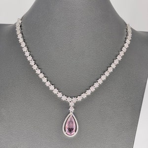 May include: A silver necklace with a teardrop-shaped pendant featuring a large pink gemstone surrounded by smaller clear gemstones.