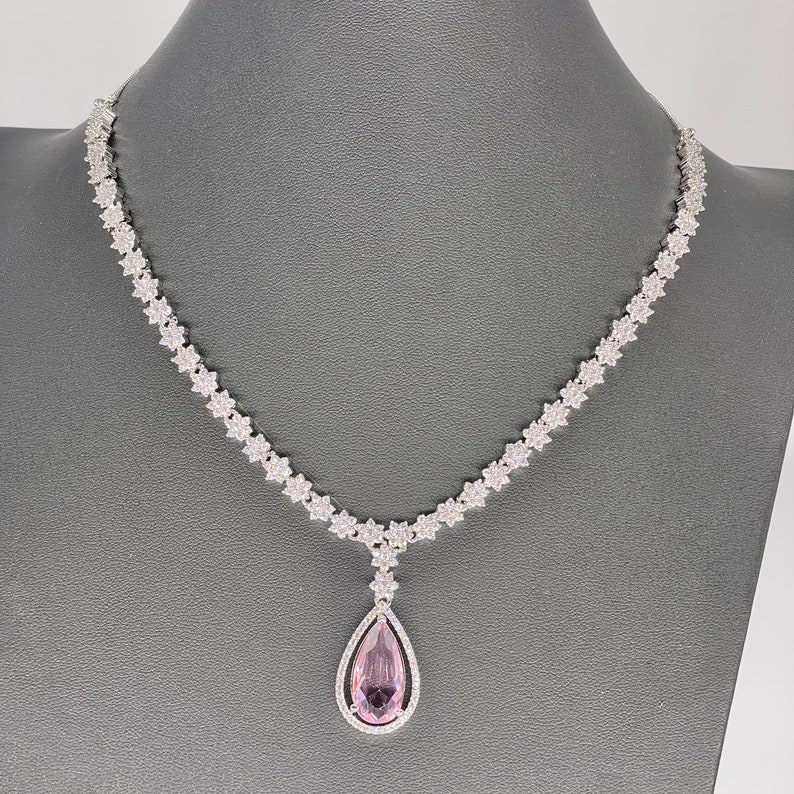 May include: A silver necklace with a teardrop-shaped pendant featuring a large pink gemstone surrounded by smaller clear gemstones.