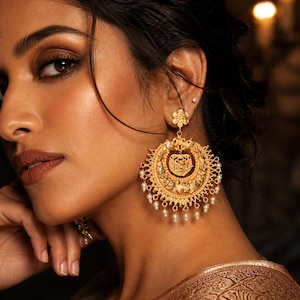 Op de afbeelding: Close-up van een vrouw met grote, sierlijke gouden oorbellen. De oorbellen zijn cirkelvormig met ingewikkelde details, waaronder een centraal ontwerp en bungelende parels. De huidskleur van de vrouw is warm en haar make-up past bij de sieraden.