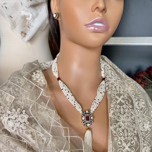 May include: A white pearl necklace with a red and gold pendant. The pendant is square shaped with a red stone in the centre and a tassel hanging from the bottom.