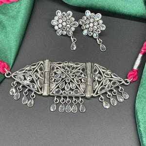 May include: A silver choker necklace with a geometric design and clear stones. The necklace has a dangling drop design. The necklace is paired with matching earrings with a flower design and clear stones.