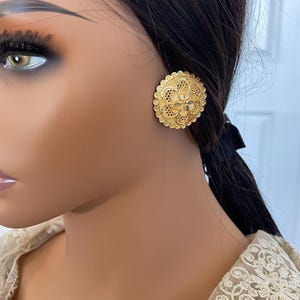 May include: A pair of gold-toned filigree earrings with a circular design. The earrings are ornate and have a delicate, intricate pattern.