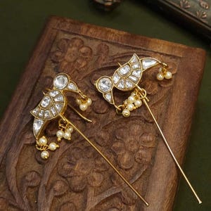 Pode incluir: Um par de brincos de ouro com um design de pássaro. Os pássaros são adornados com pedras brancas e têm uma corrente de pérolas brancas penduradas.
