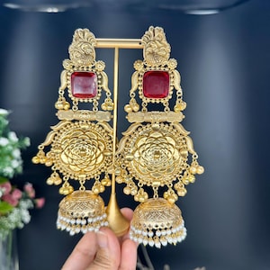 May include: Gold-coloured earrings with a red square gemstone at the top. The earrings feature intricate floral and geometric designs, with small dangling beads and a bell-shaped bottom adorned with pearls. The earrings are displayed on a gold stand.
