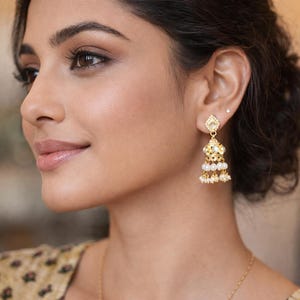 Op de afbeelding: Goudkleurige oorhangers met een diamantvormige bovenkant en drie rijen witte parels. De oorbellen worden gedragen op het oor van een persoon, met een zachte achtergrond.