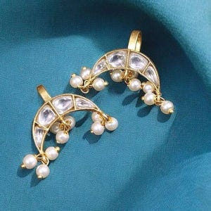 May include: A pair of gold crescent-shaped nose rings with white pearl accents. The rings are decorated with clear stones.