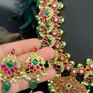 May include: A gold necklace and earrings set with pink and green gemstones. The necklace features a geometric design with multiple layers of stones. The earrings are teardrop shaped with a similar design.