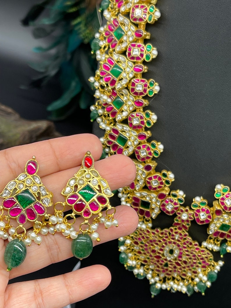 May include: A gold necklace and earrings set with pink and green gemstones. The necklace features a geometric design with multiple layers of stones. The earrings are teardrop shaped with a similar design.