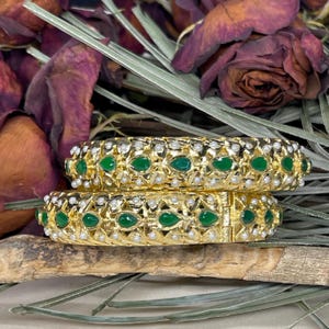 Pode incluir: Dois braceletes de ouro com pedras verdes e pérolas brancas. Os braceletes estão empilhados um em cima do outro.
