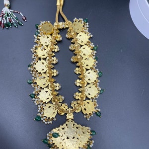 May include: A gold necklace with a large, ornate pendant and green and white beads. The pendant is shaped like a flower and has intricate details.