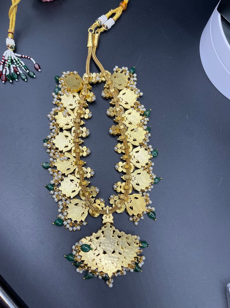 May include: A gold necklace with a large, ornate pendant and green and white beads. The pendant is shaped like a flower and has intricate details.