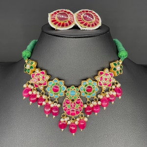 May include: A gold necklace with a green, pink, and blue floral design. The necklace has a pendant with a crescent moon shape and is adorned with red and white beads. The necklace is paired with matching earrings.