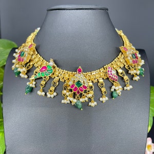 May include: A gold-toned necklace with intricate detailing, featuring red, green, and white accents. The necklace has a series of pendant drops with pearls and emerald-colored stones. The design includes floral and bird-like motifs.