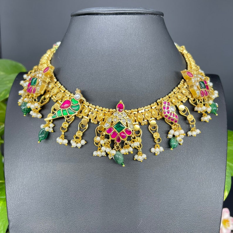 May include: A gold-toned necklace with intricate detailing, featuring red, green, and white accents. The necklace has a series of pendant drops with pearls and emerald-colored stones. The design includes floral and bird-like motifs.