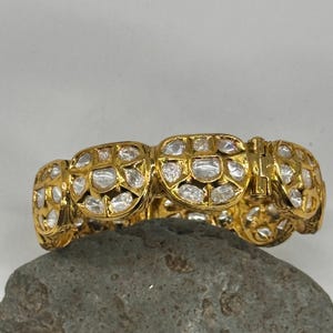 May include: A gold hinged bangle bracelet featuring a series of interconnected circular and oval links. Each link is set with multiple clear, flat-cut stones arranged in a geometric pattern. The metal has a textured, warm yellow gold finish.