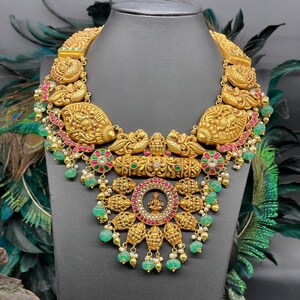 May include: A gold necklace with intricate detailing and green and pink gemstones. The necklace features a central pendant with a circular design and a small figure in the centre.
