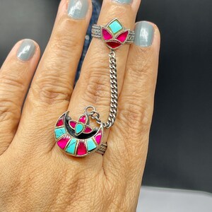 May include: A silver ring with a turquoise and pink enamel design in the shape of a crescent moon. The ring is connected to a smaller ring with a flower design by a silver chain.