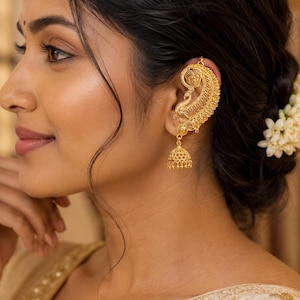 May include: A gold-toned ear cuff featuring an intricate filigree design that follows the curve of the ear, accented with a small, bell-shaped jhumka dangle hanging from the lower portion. The metalwork displays detailed patterns and textures.