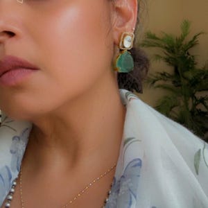 May include: Gold-toned earrings with a two-tiered design. The top tier is a square shape with a white stone, and the bottom tier is a teardrop-shaped, green stone. The earrings are worn on the ear of a person wearing a floral patterned scarf.