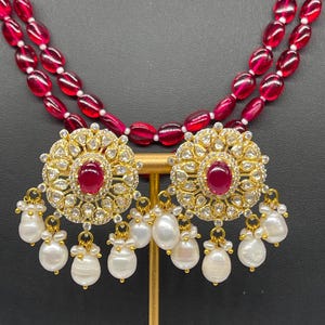 Könnte beinhalten: Eine zweireihige Halskette aus roten Perlen mit einem Paar goldenen Ohrringen, die einen großen roten Stein umgeben von kleineren weißen Steinen und Perlentropfen aufweisen.