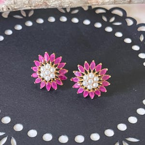 May include: A pair of gold stud earrings featuring a pink flower design. The centre of each flower is decorated with a cluster of white pearls.