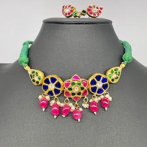 Op de afbeelding: Een goudkleurige ketting- en oorbellenset. De ketting heeft blauwe en roze bloemmotieven met bungelende roze kralen en kleine witte parels. De oorbellen hebben een vergelijkbaar ontwerp met rode en groene accenten. De ketting zit op een groen koord.