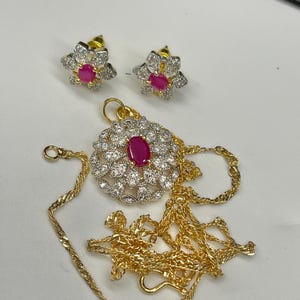 May include: A gold chain necklace with a pendant featuring a round, floral design with a pink gemstone in the center. The pendant is surrounded by smaller white gemstones. The set also includes a pair of matching earrings with a similar floral design and pink gemstones.