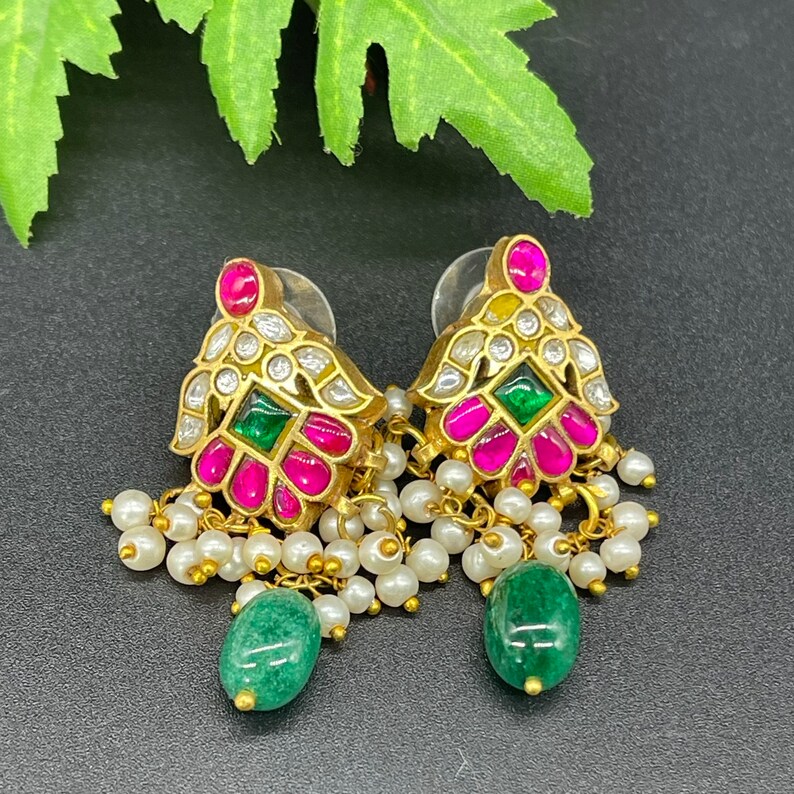 May include: Gold-toned earrings with a floral design, featuring pink and green gemstones, and small white pearls. The earrings have a dangling green stone at the bottom. The earrings are set against a black background.