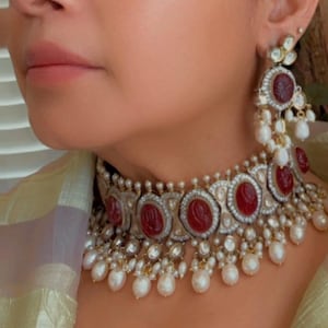 May include: Close-up of a luxurious jewelry set featuring a necklace and earrings. The necklace and earrings have red gemstone centerpieces surrounded by small white stones and dangling white pearls. The jewelry is set against a soft, neutral background.