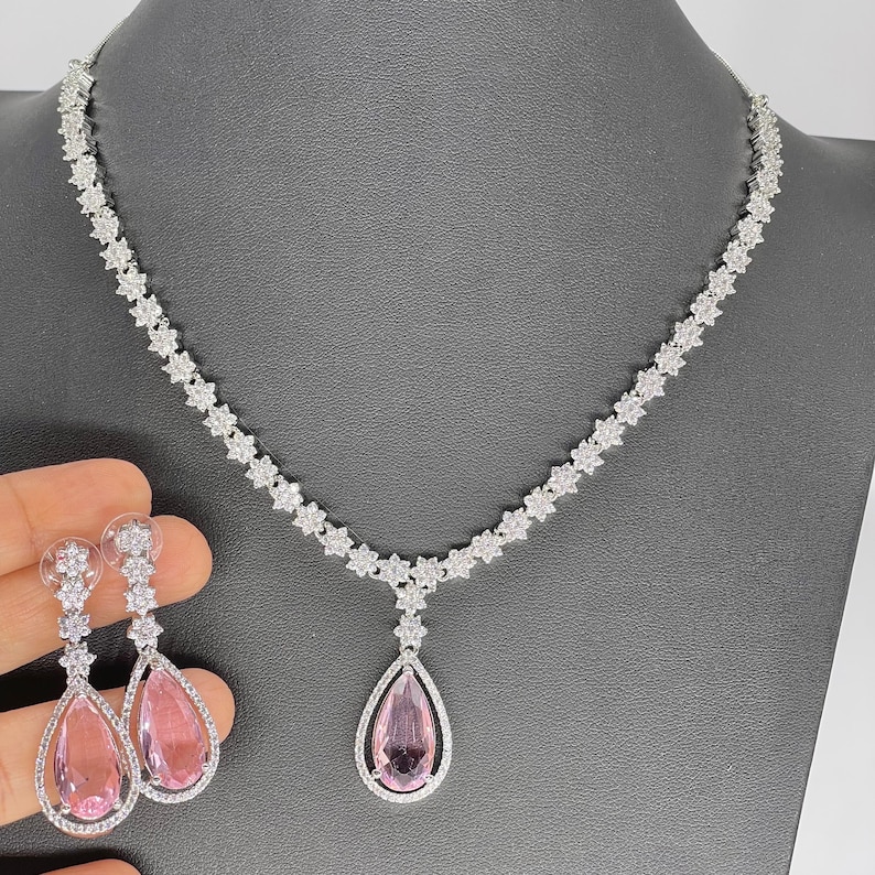 May include: A silver necklace with a teardrop-shaped pendant featuring a pink crystal. The necklace is adorned with small, clear crystals in a floral pattern. Matching earrings with pink crystals are also included.