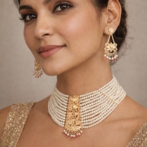 Op de afbeelding: Een vrouw draagt een gouden ketting en oorbellen. De ketting is een meerrijige parelchoker met een gouden hanger. De oorbellen zijn goudkleurig met parel- en rode kraalaccenten. De vrouw draagt een gouden pailletten top.