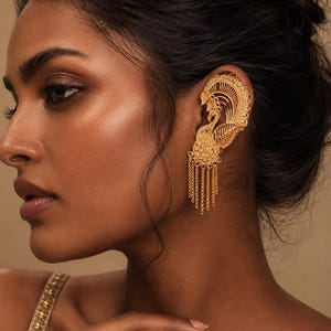 Peut inclure: Un ear cuff doré avec un motif de paon. Ce bijou ornemental présente une silhouette de paon avec une queue en éventail et des chaînes pendantes. Le ear cuff est porté sur l'oreille et est visible contre la peau.