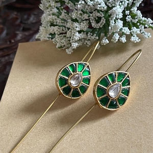 Peut inclure: Une paire de boucles d'oreilles dorées avec un motif en forme de goutte d'eau. Chaque boucle d'oreille présente une pierre transparente centrale entourée de pierres vertes facettées. Les boucles d'oreilles ont de longues tiges fines.