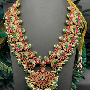 May include: A gold necklace with pink and green gemstones. The necklace has a pendant in the shape of a flower with a pearl and green gemstone drop.