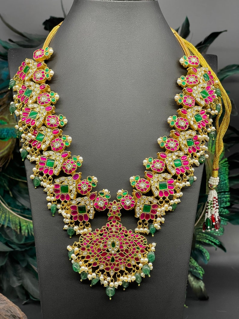 May include: A gold necklace with pink and green gemstones. The necklace has a pendant in the shape of a flower with a pearl and green gemstone drop.