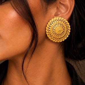 Peut inclure: Une grande boucle d'oreille à clou dorée et circulaire, présentant un motif complexe de style filigrane avec des anneaux concentriques de détails métalliques texturés. Le design inclut un motif floral central entouré de couches festonnées.