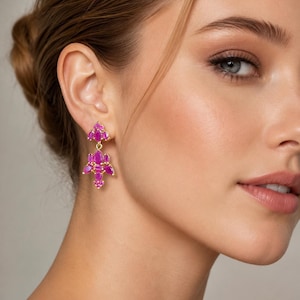 Könnte beinhalten: Goldfarbene Ohrhänger mit fuchsiafarbenen Edelsteinen. Die Ohrringe haben ein florales Design, mit mehreren symmetrisch angeordneten Edelsteinen. Die Ohrringe werden am Ohr einer Person getragen.