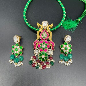 May include: A gold necklace with a peacock pendant featuring green, pink, and white stones. The necklace is paired with matching earrings with green, pink, and white stones.