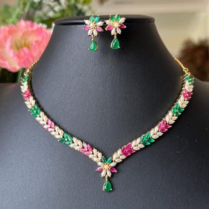 May include: A gold-toned necklace and earring set. The necklace features a leaf design with pink, green, and white stones. The earrings have a flower design with matching stones and a green teardrop. The jewelry is displayed on a black mannequin.