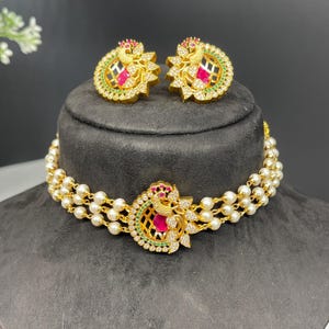 May include: A gold necklace with a three-strand pearl choker and a peacock pendant with red and green stones. Matching earrings with peacock designs and red and green stones.