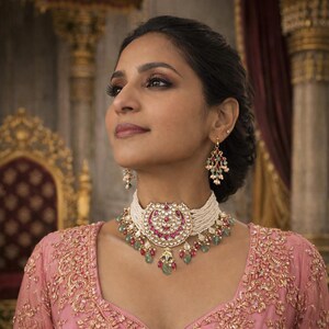 Op de afbeelding: Een vrouw draagt een choker ketting en bijpassende oorbellen. De ketting heeft een grote, ronde hanger met rode en witte stenen, omringd door kleinere groene stenen. De oorbellen hebben een vergelijkbaar ontwerp. De vrouw draagt een roze geborduurd topje.