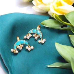 May include: Two gold crescent-shaped nose rings with turquoise inlay and pearl accents. The rings are on a teal fabric background.