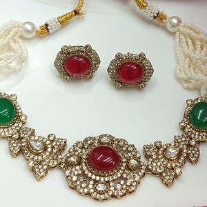 May include: A gold necklace with a white pearl strand and a center pendant featuring red and green gemstones surrounded by clear rhinestones. Matching earrings with red gemstones are also included.