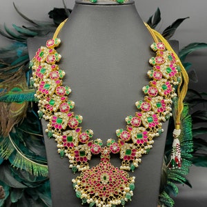 May include: A gold necklace with pink and green gemstones, a matching pair of earrings, and a pearl tassel. The necklace features a floral design with intricate details.