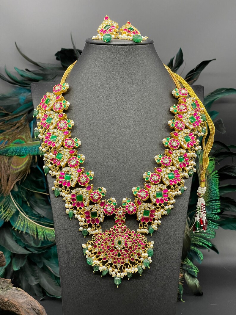 May include: A gold necklace with pink and green gemstones, a matching pair of earrings, and a pearl tassel. The necklace features a floral design with intricate details.