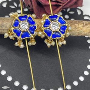 May include: A pair of gold earrings with blue enamel flower designs and white pearl accents. Each earring features a clear rhinestone in the center of the flower.