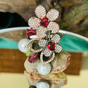 May include: A silver ring with a cluster of three flowers. The flowers are made of white and black stones with red accents. The ring is set on a rock with a reflective surface.