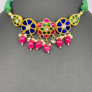 Op de afbeelding: Een goudkleurige ketting met een groene koord. De ketting heeft blauwe en roze bloemmotieven met witte accenten en bungelende roze kralen. De ketting staat tegen een donkergrijze achtergrond.