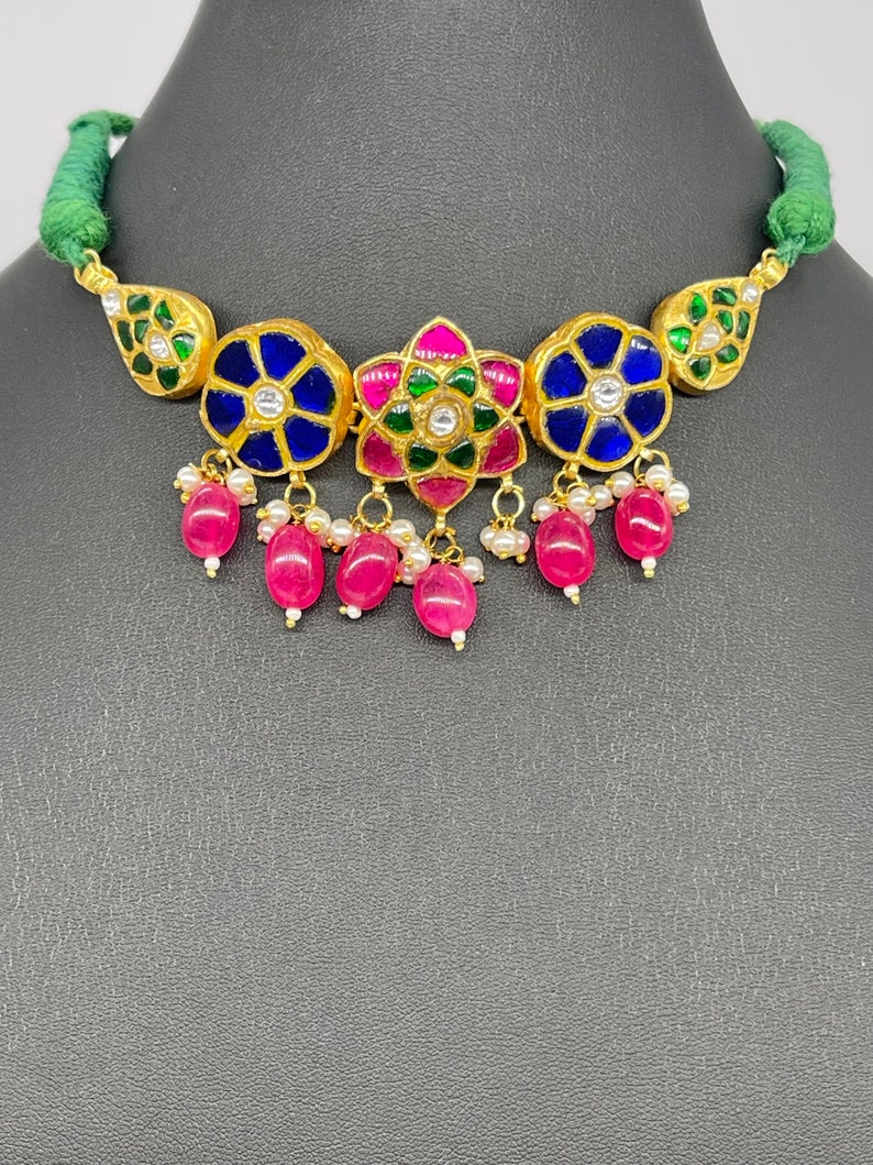 Op de afbeelding: Een goudkleurige ketting met een groene koord. De ketting heeft blauwe en roze bloemmotieven met witte accenten en bungelende roze kralen. De ketting staat tegen een donkergrijze achtergrond.