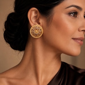 May include: A close-up of a gold-colored, round earring with an intricate, openwork design. The earring is a stud style, featuring a central floral element surrounded by a detailed pattern and small beaded edge. The earring is worn on a person's ear.